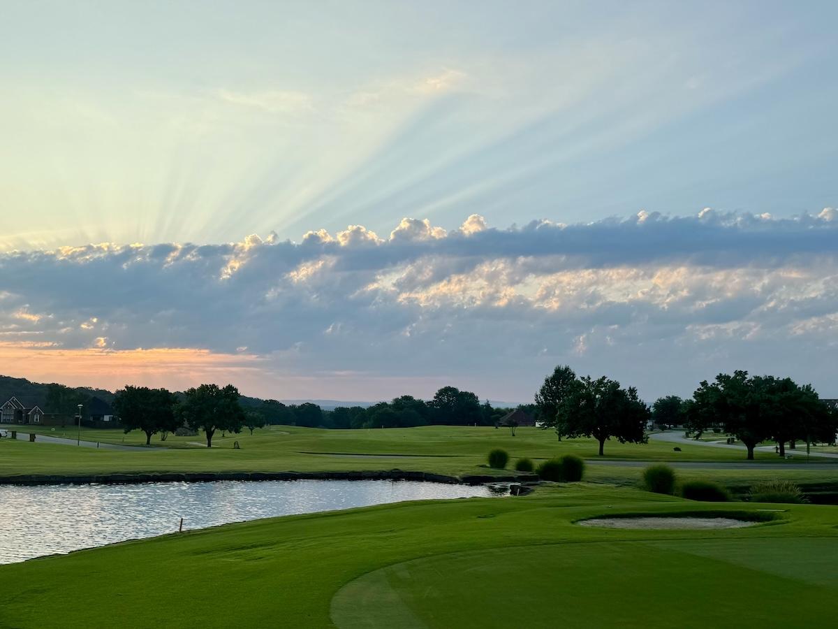 Sunset over a golf course with a pond and trees, rays of light through clouds.