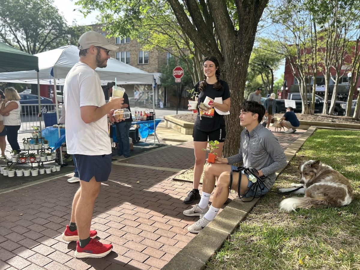 Three people chat under a tree on a sunny day, with drinks. A dog sits nearby.