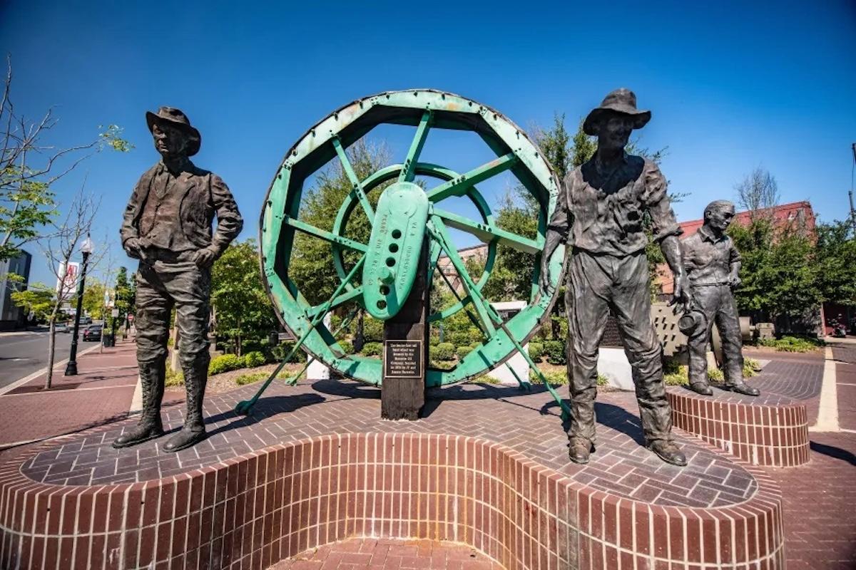 Bronze statues of workers near a large green wheel outdoors.