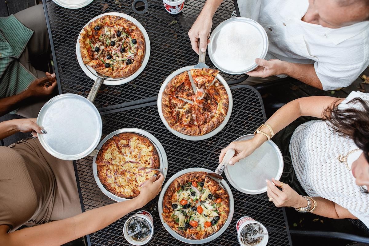 Four people sharing five pizzas at a table outdoors.