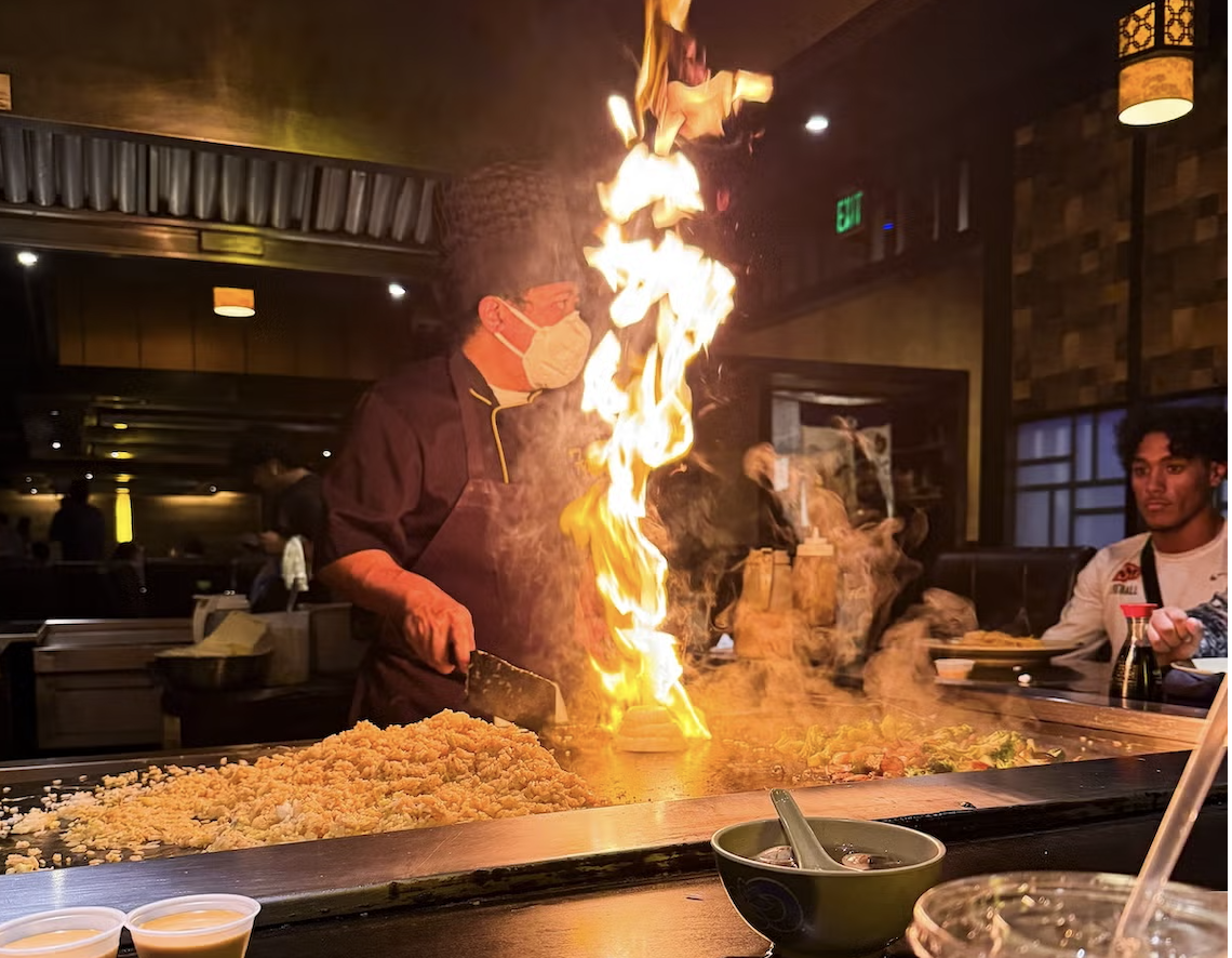 Chef cooking with a large flame on a hibachi grill in a dimly lit restaurant.