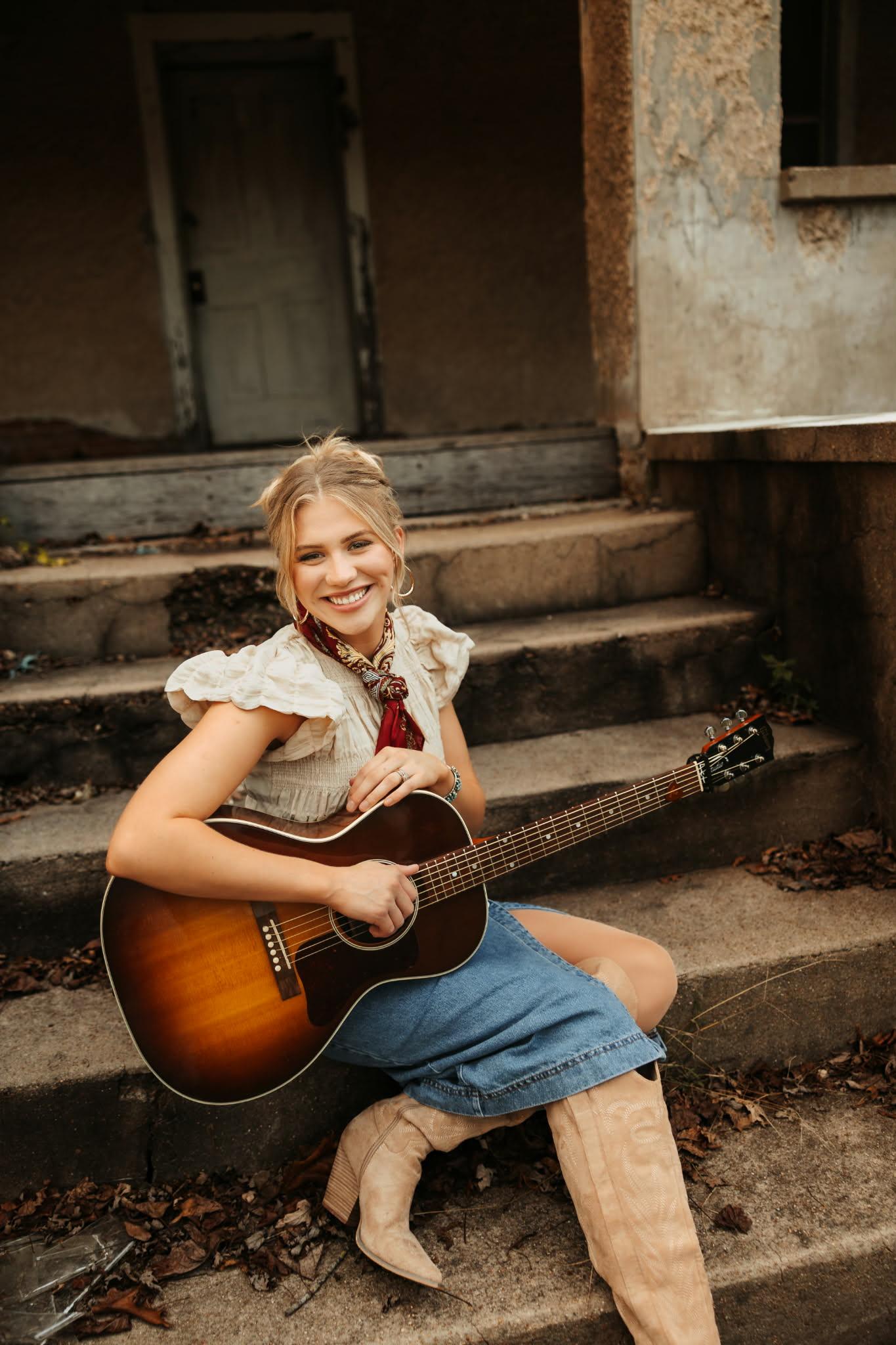 Hannah Harper sitting on steps, smiling, holding a guitar, wearing boots and a skirt.