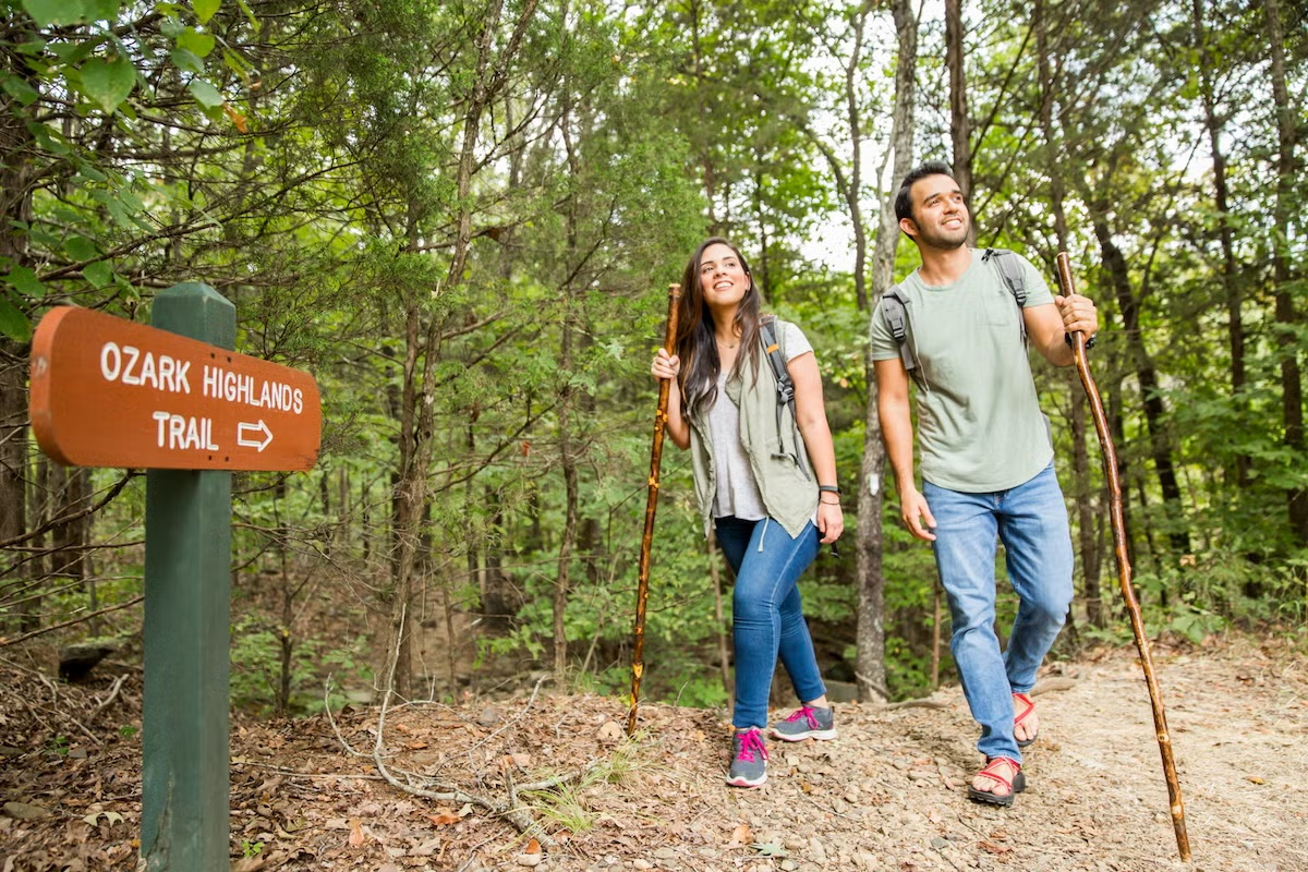 Two people hiking on a forest trail, smiling and holding walking sticks.