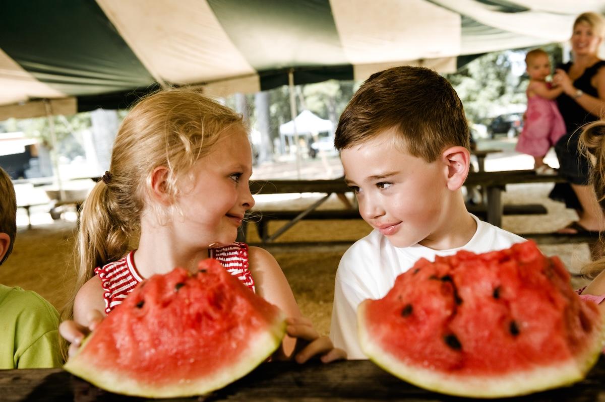 Children smiling with watermelon slices at a picnic table.