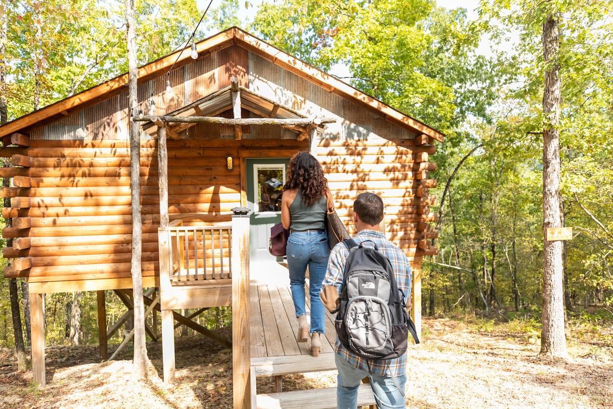 Two people with backpacks approaching a wooden cabin in the woods.