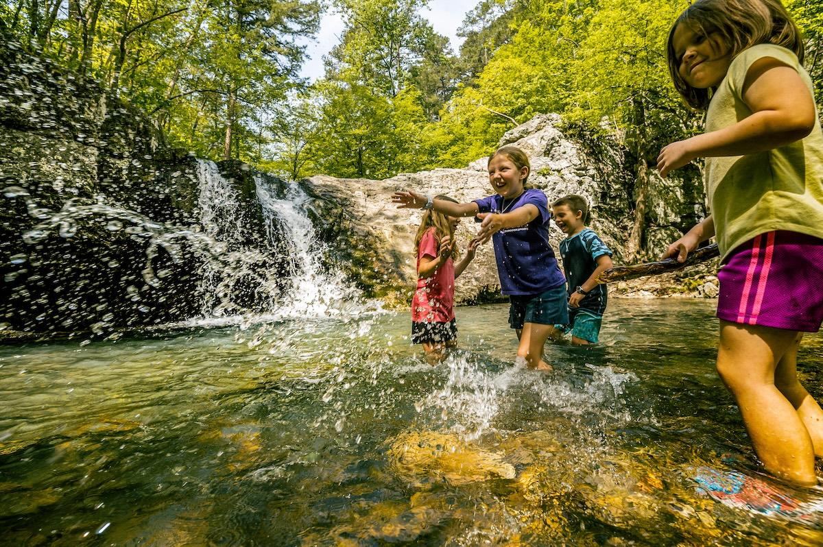 Children playing in a forest stream near a waterfall, surrounded by lush greenery.