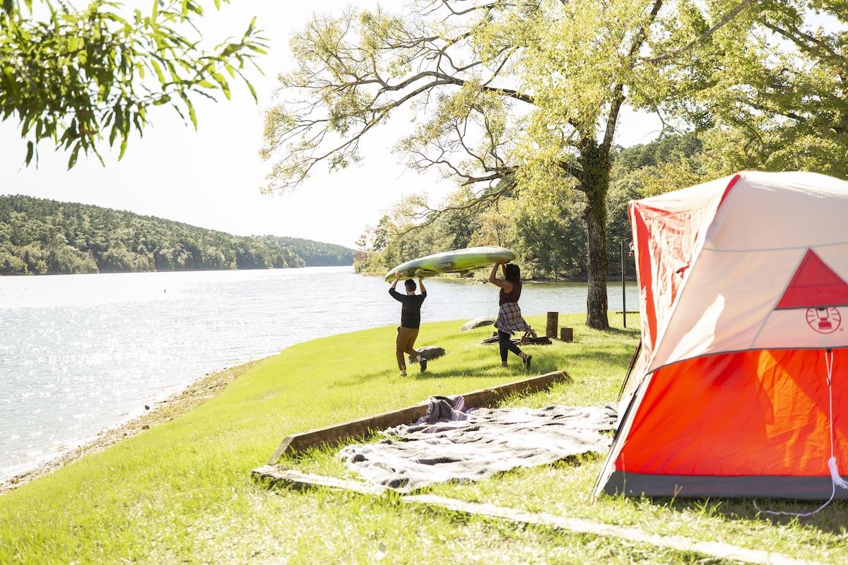 Tent by a lakeside, two people lifting a kayak under a sunny sky.