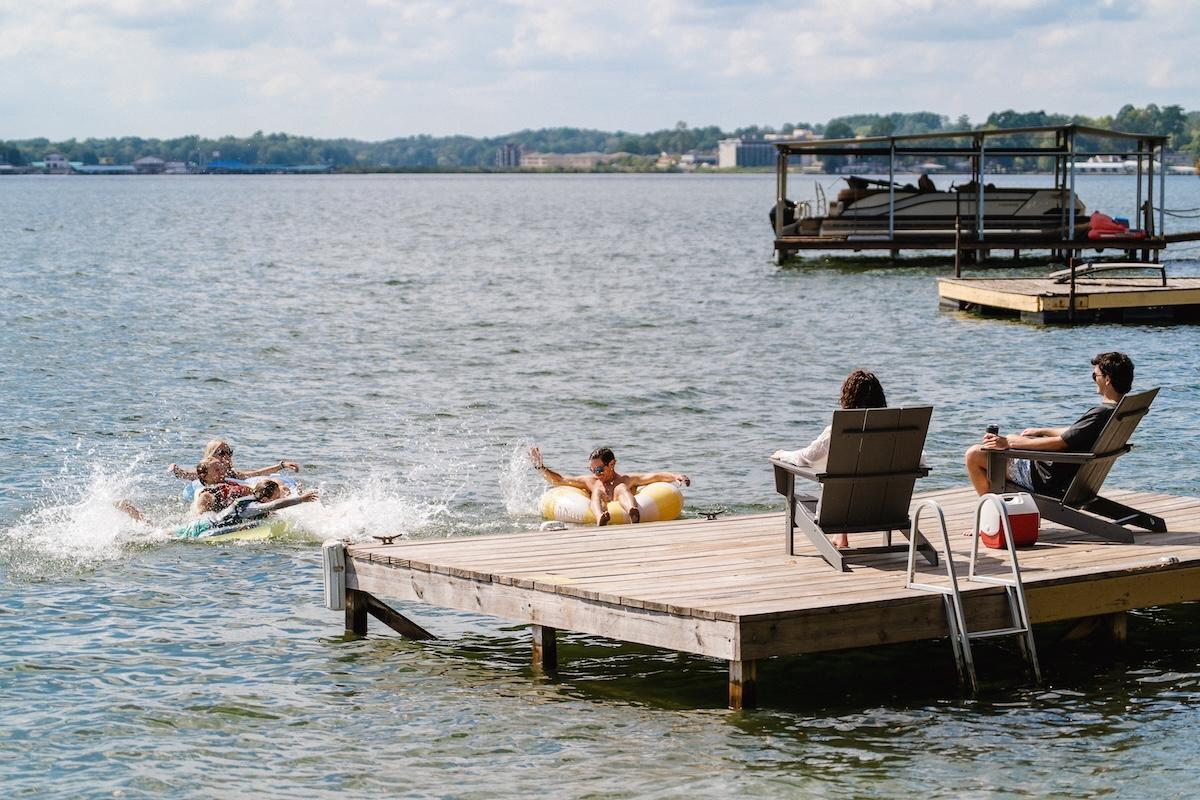 Two people float near a lakeside dock with two others seated, enjoying the sunny day.