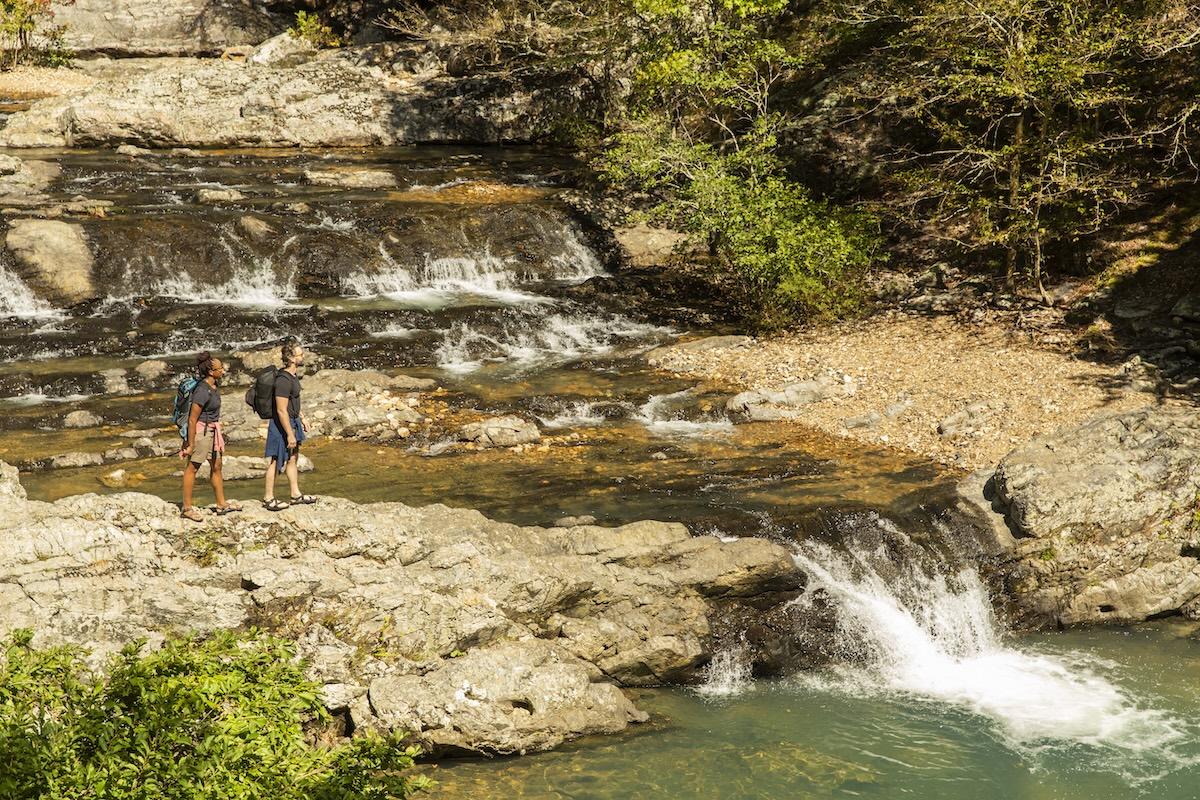 Two people hike near a rocky waterfall, surrounded by greenery.