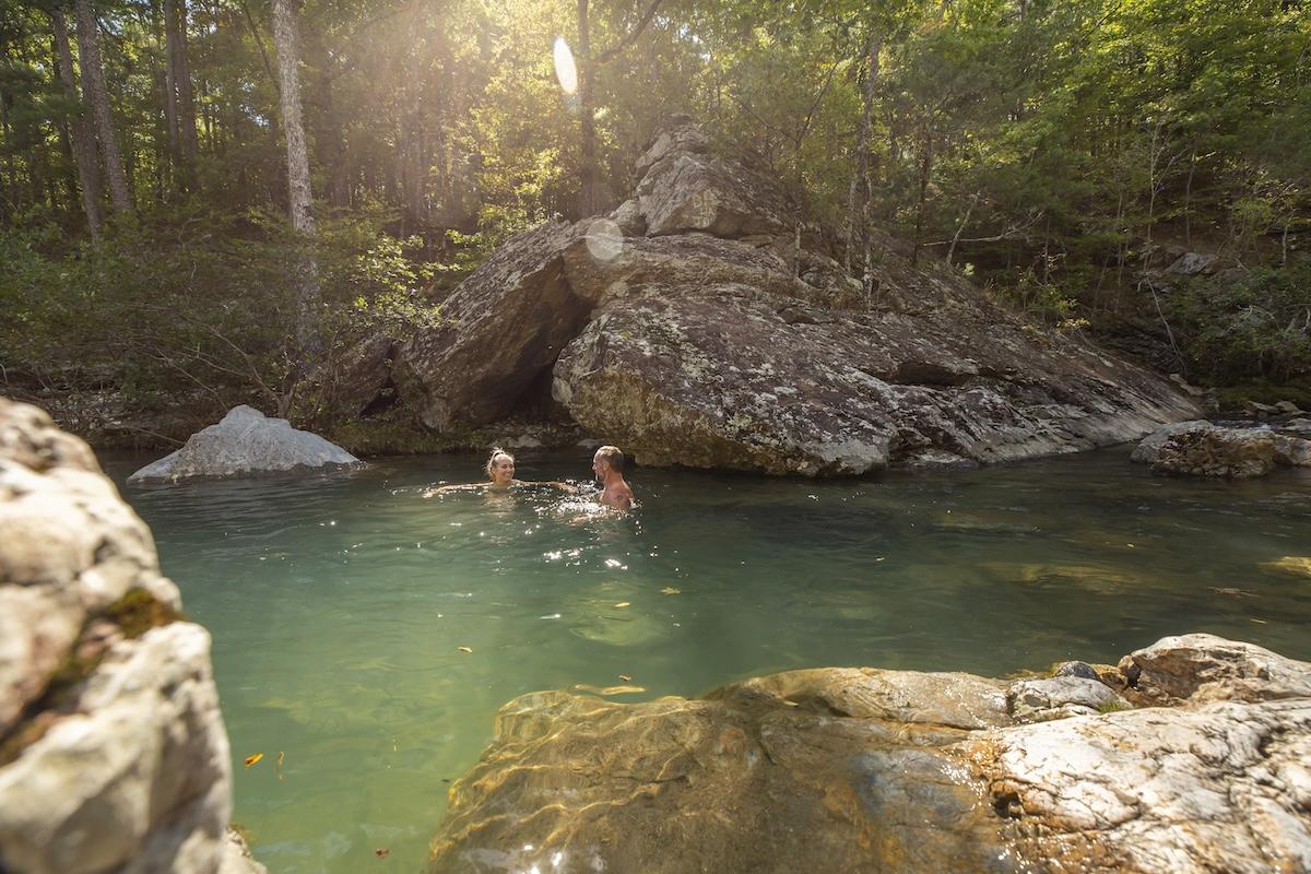 Two people swimming in a sunlit forest river, surrounded by rocks and trees.