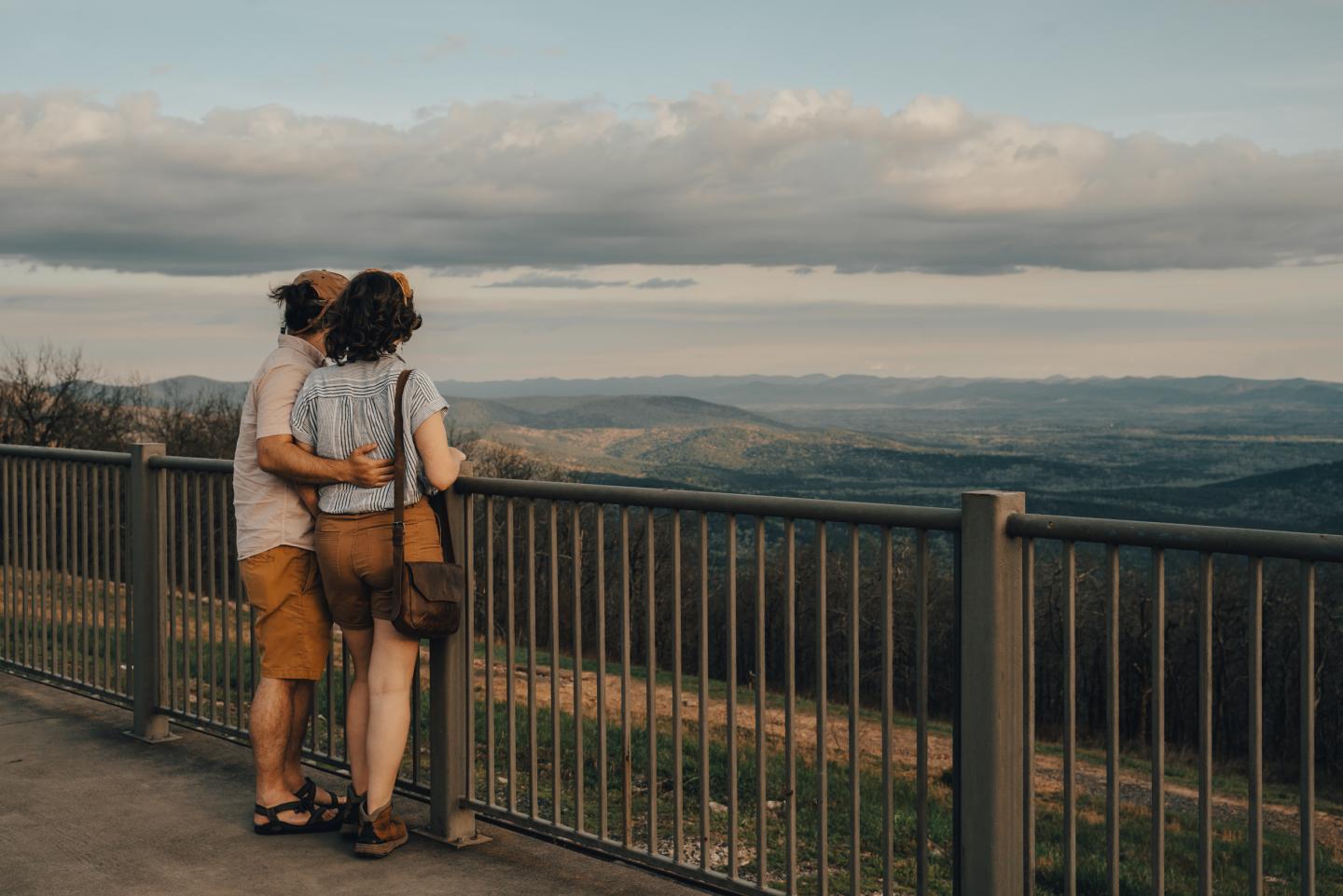 Couple embracing at scenic overlook, viewing mountains under cloudy sky.