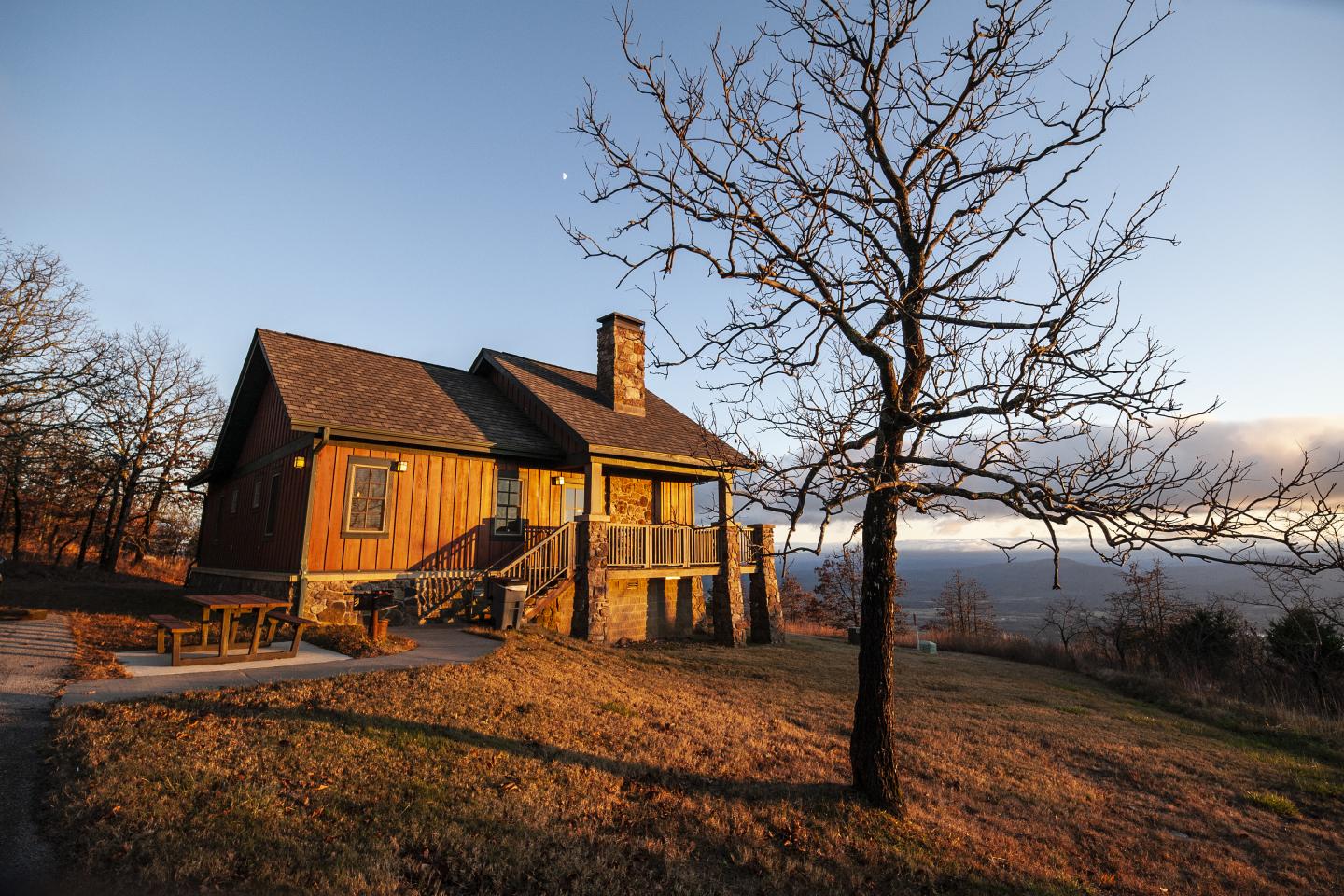 Cozy cabin in a grassy field at sunset, with a bare tree nearby.