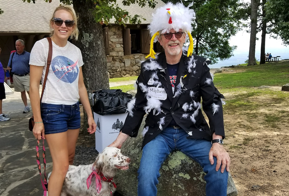 Woman with dog next to man in a feathered costume, outdoors.