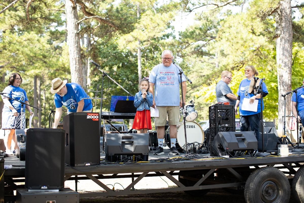People on an outdoor stage, speaking into microphones, surrounded by trees.