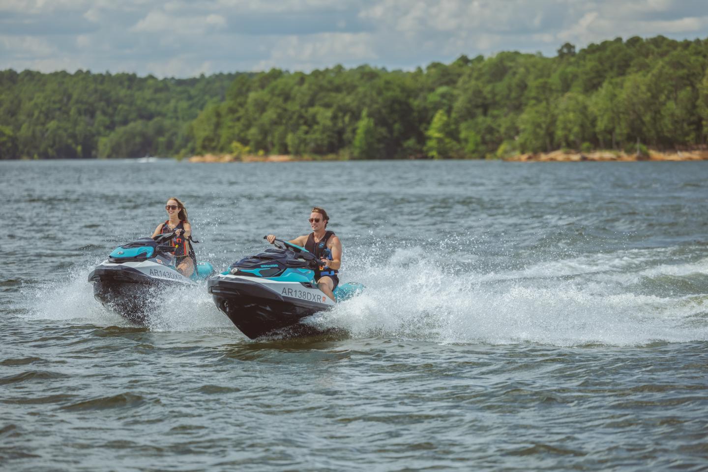 Two people riding jet skis on a lake, surrounded by trees.
