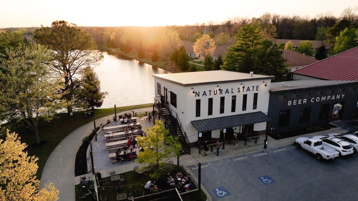 Brewery by a lake at sunset with outdoor seating and parked cars.