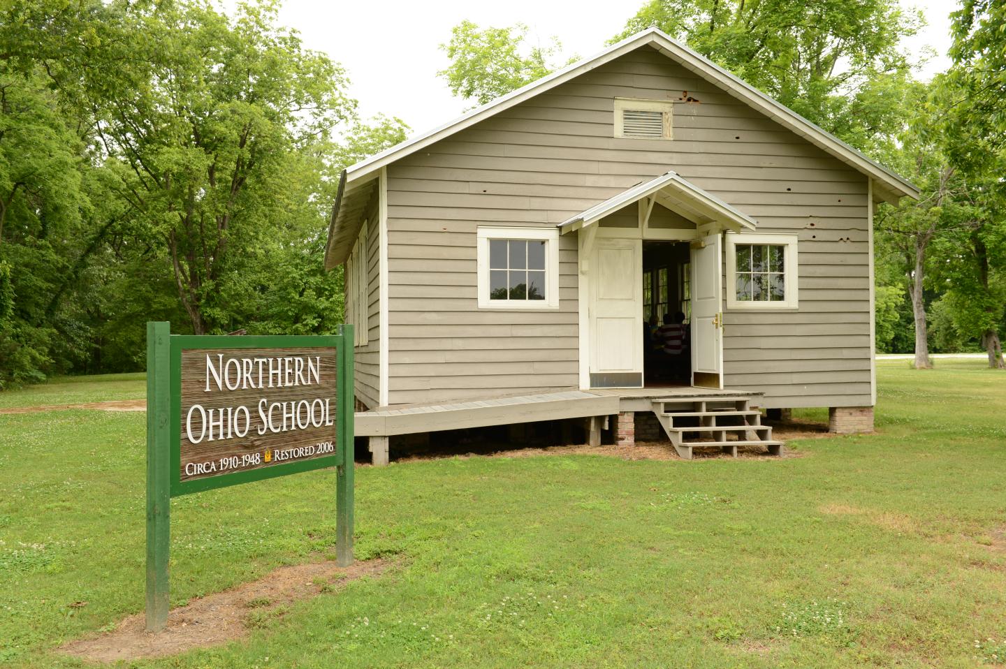 Small wooden schoolhouse with a sign, surrounded by trees.