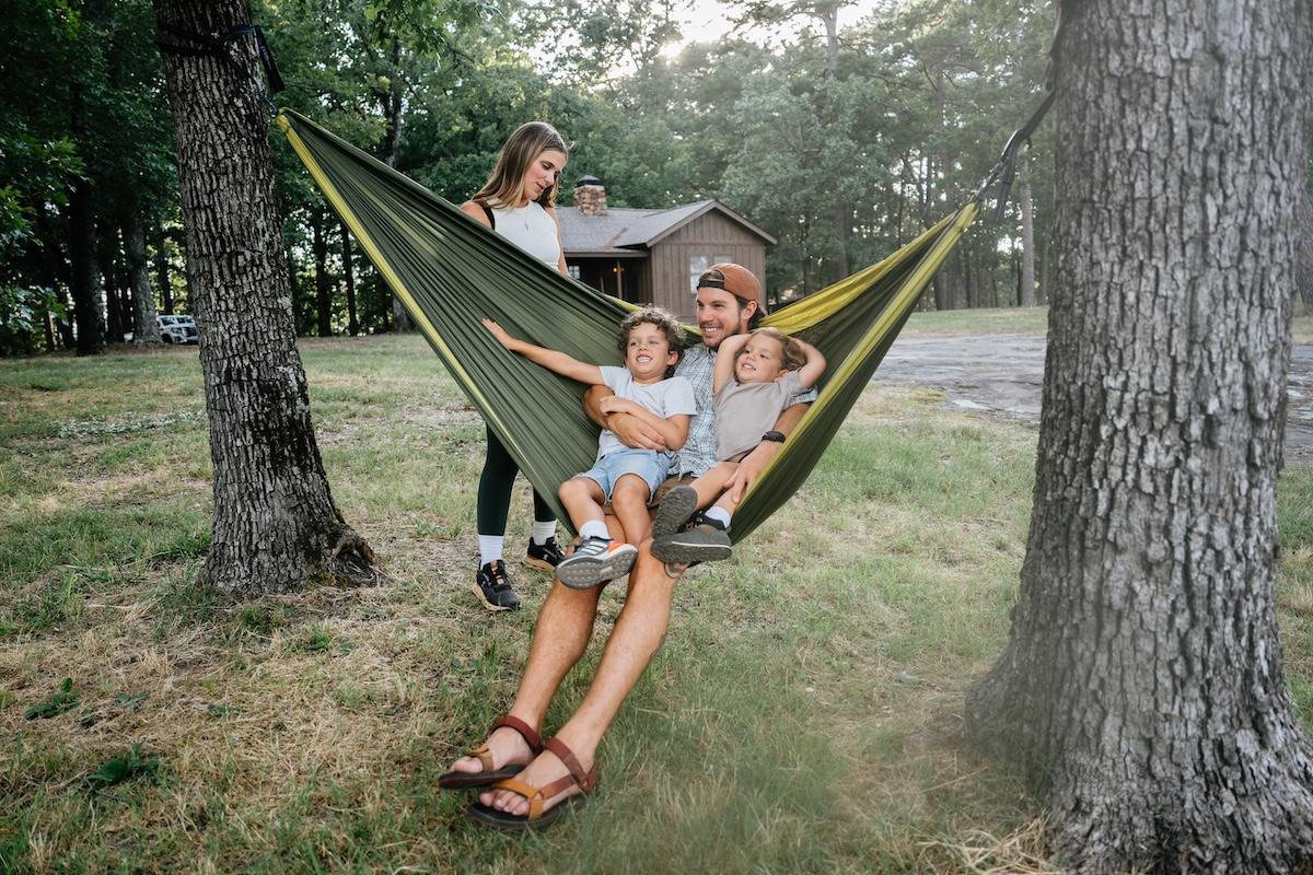 Family relaxing in a hammock between trees, with a cabin in the background.