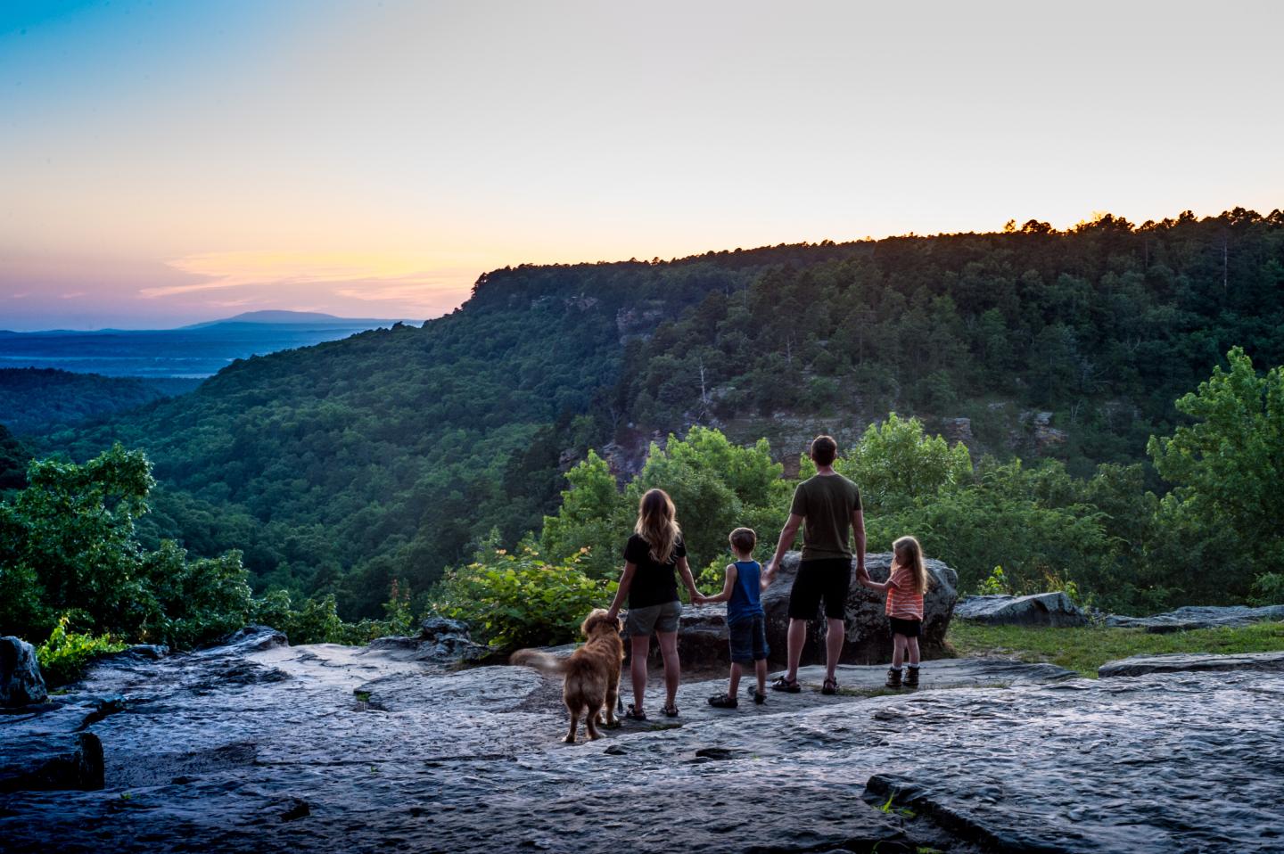 Family with dog on rocky overlook enjoying sunset view over forested hills.