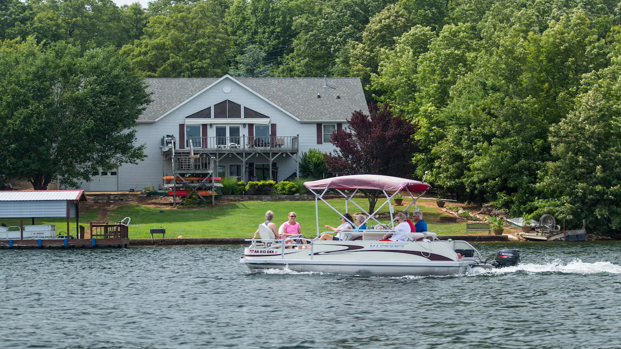 Pontoon boat with people, near a lakeside house, surrounded by trees.