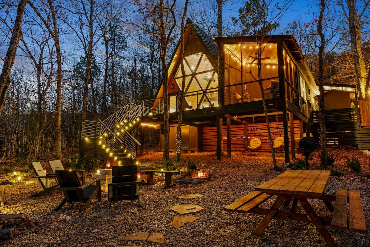 A-frame cabin in woods with warm lights at dusk.