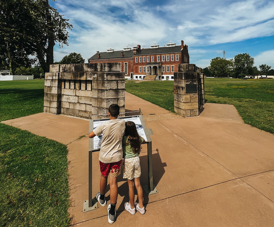 Two children look at an outdoor exhibit near a historic building.