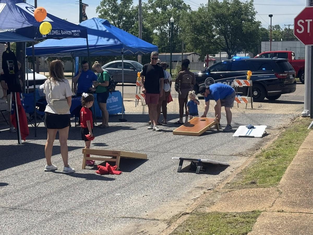 Street fair with people playing cornhole under blue tents.