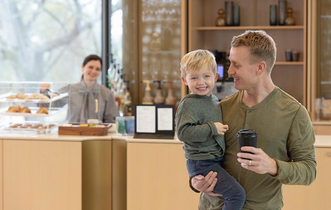 Man holding a smiling child, standing in a café, while a barista arranges pastries.