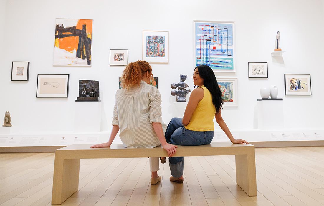 Two women sit on a bench in an art gallery, viewing paintings on the wall.