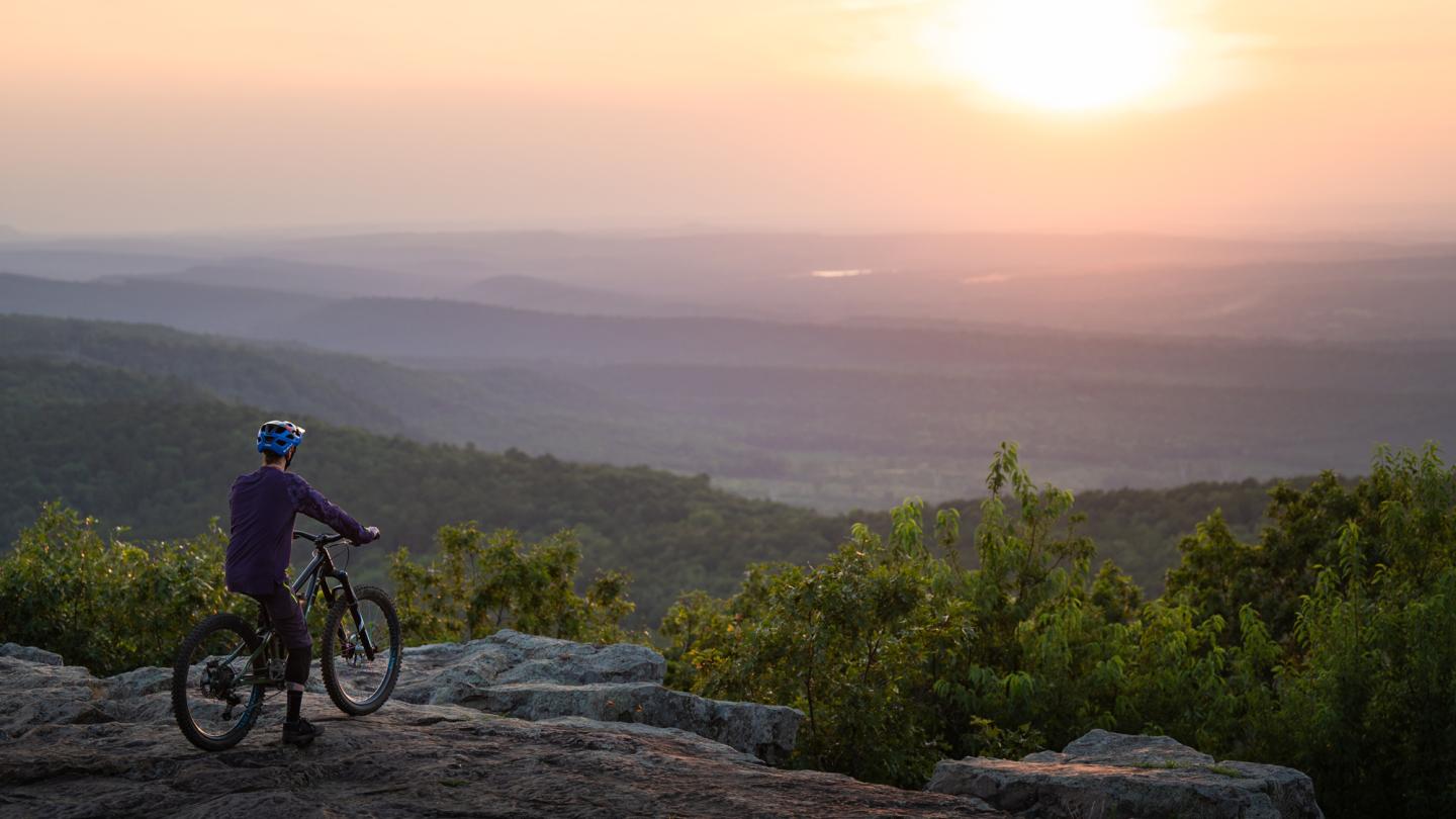 Cyclist on rocky hill at sunset overlooking a vast landscape.