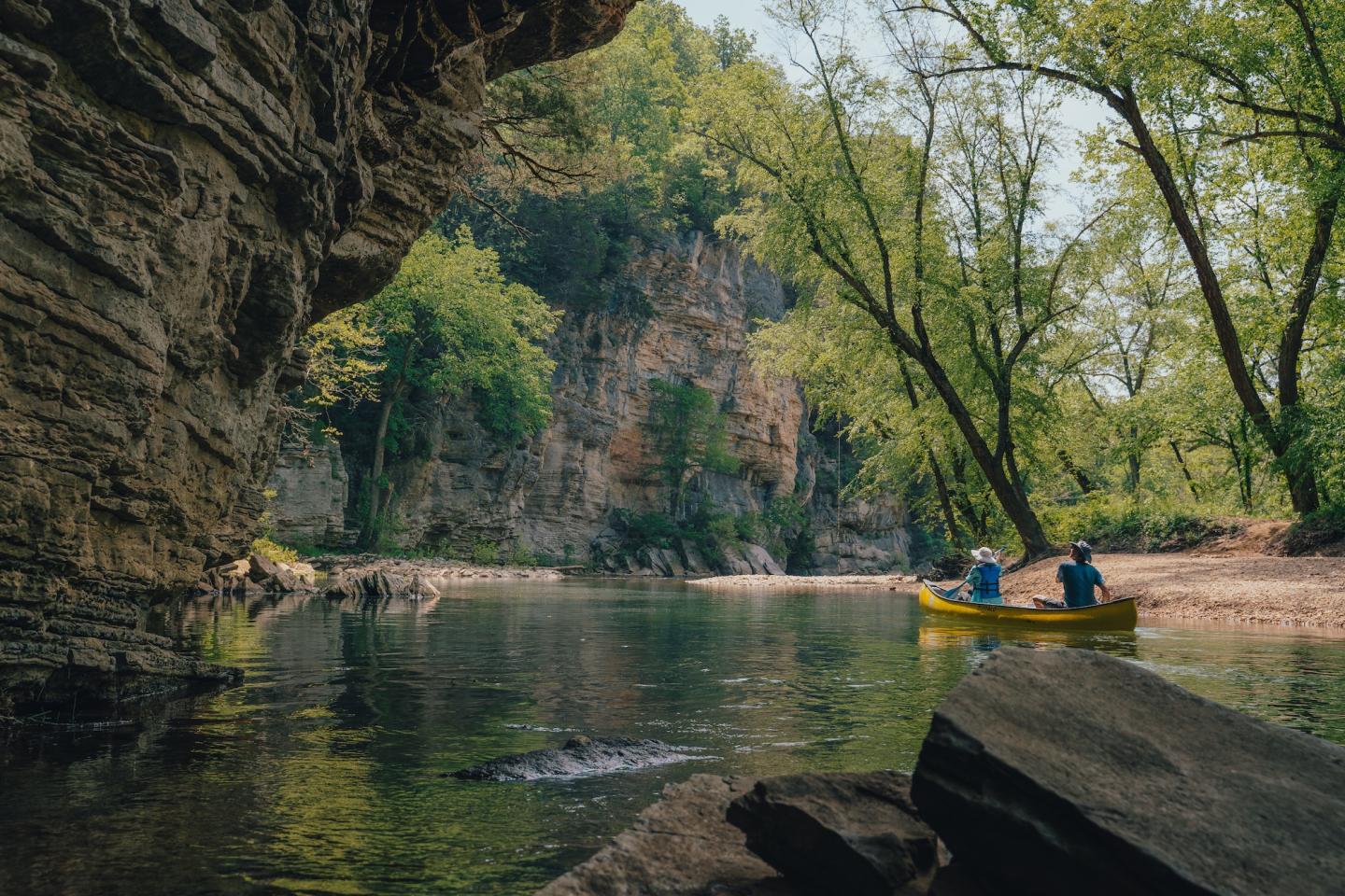 Kayakers in a yellow canoe on a calm river, surrounded by cliffs and trees.