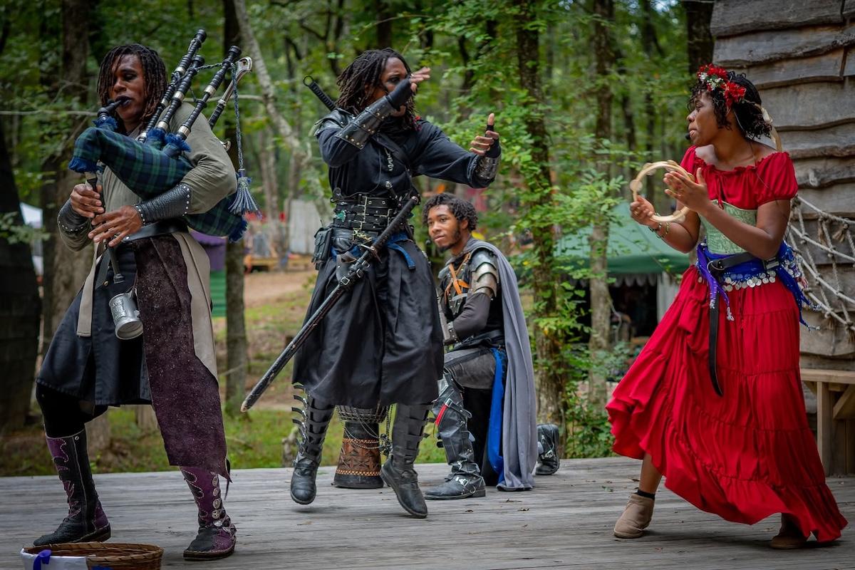 Actors in medieval costumes on stage in a forest setting, playing instruments and acting.