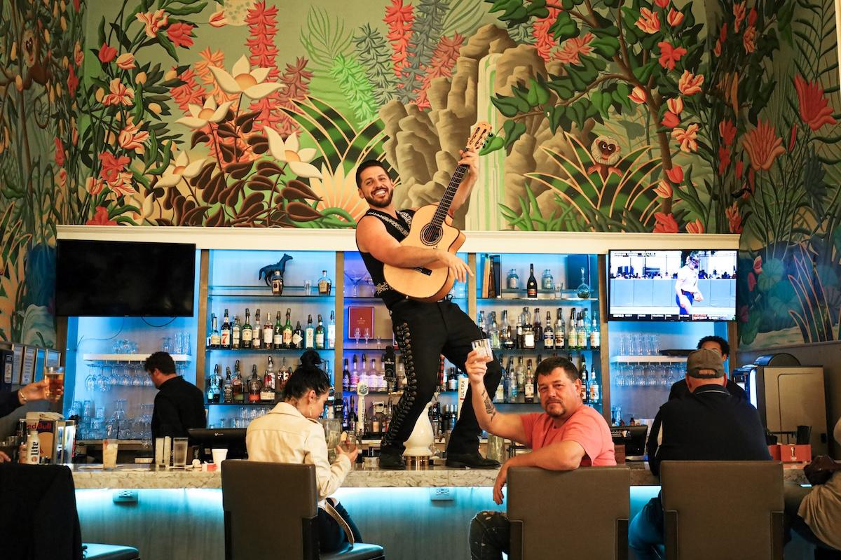Man standing on bar playing guitar, vibrant mural behind.