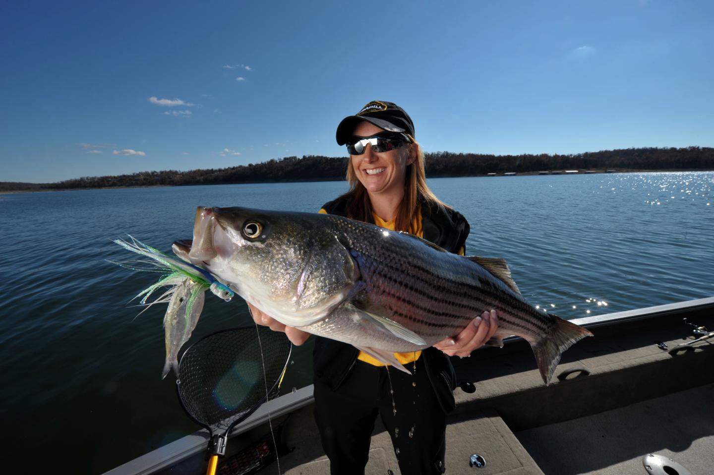 Woman smiling on a boat, holding a large fish on a sunny day.
