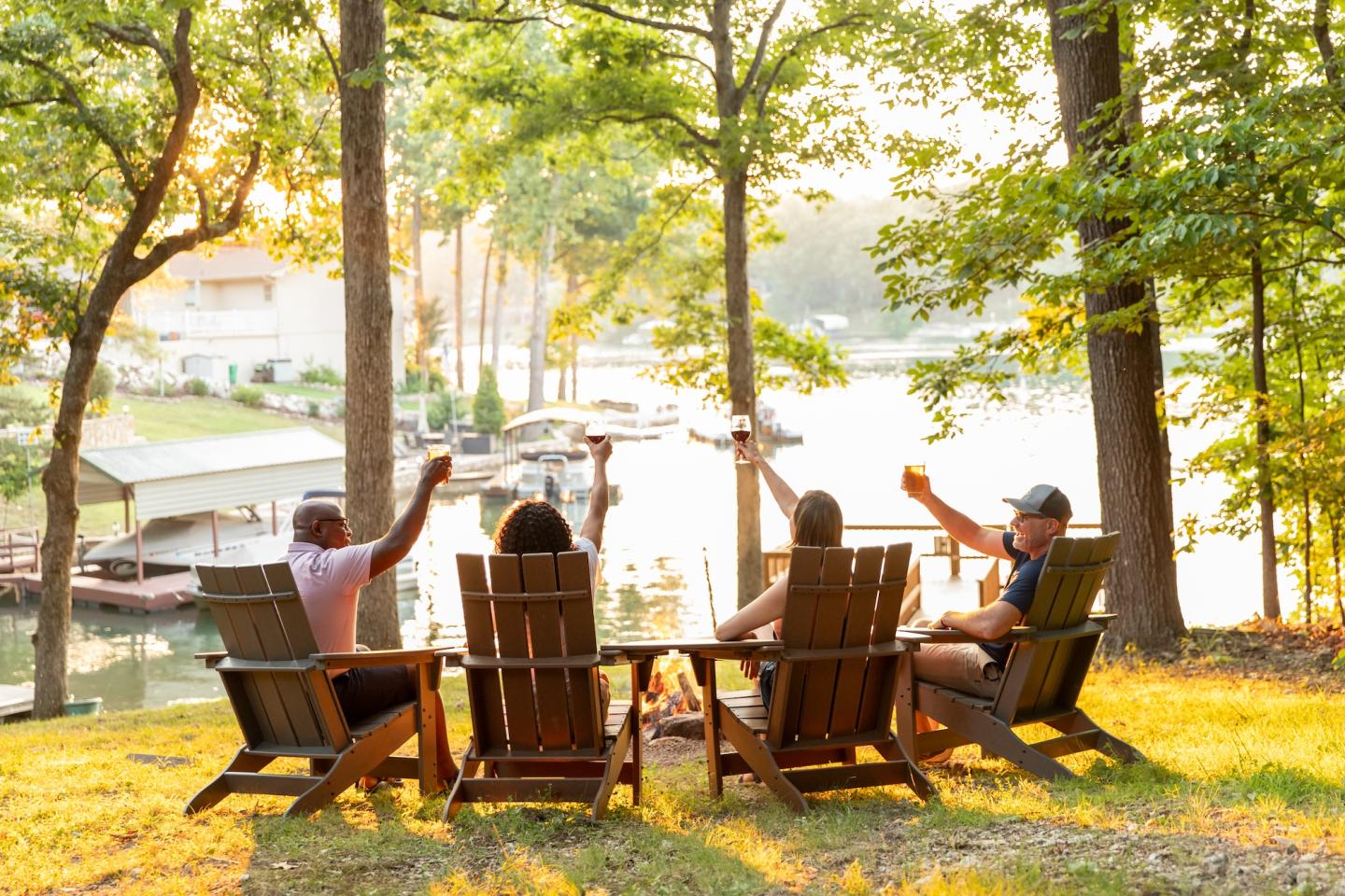 Four people in chairs toasting by a lake, surrounded by trees and docked boats.
