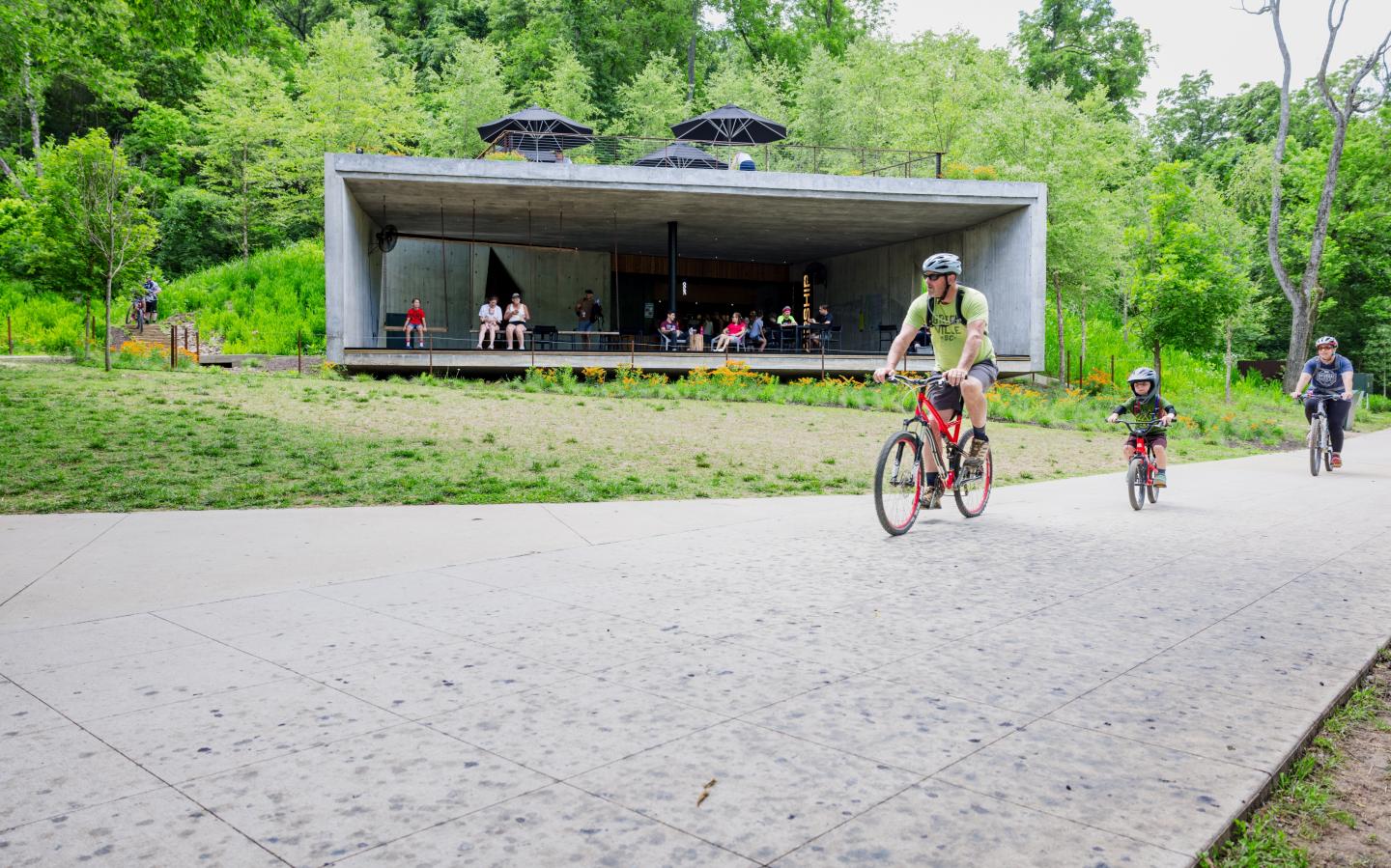 Cyclists ride past a modern pavilion in a lush park setting.