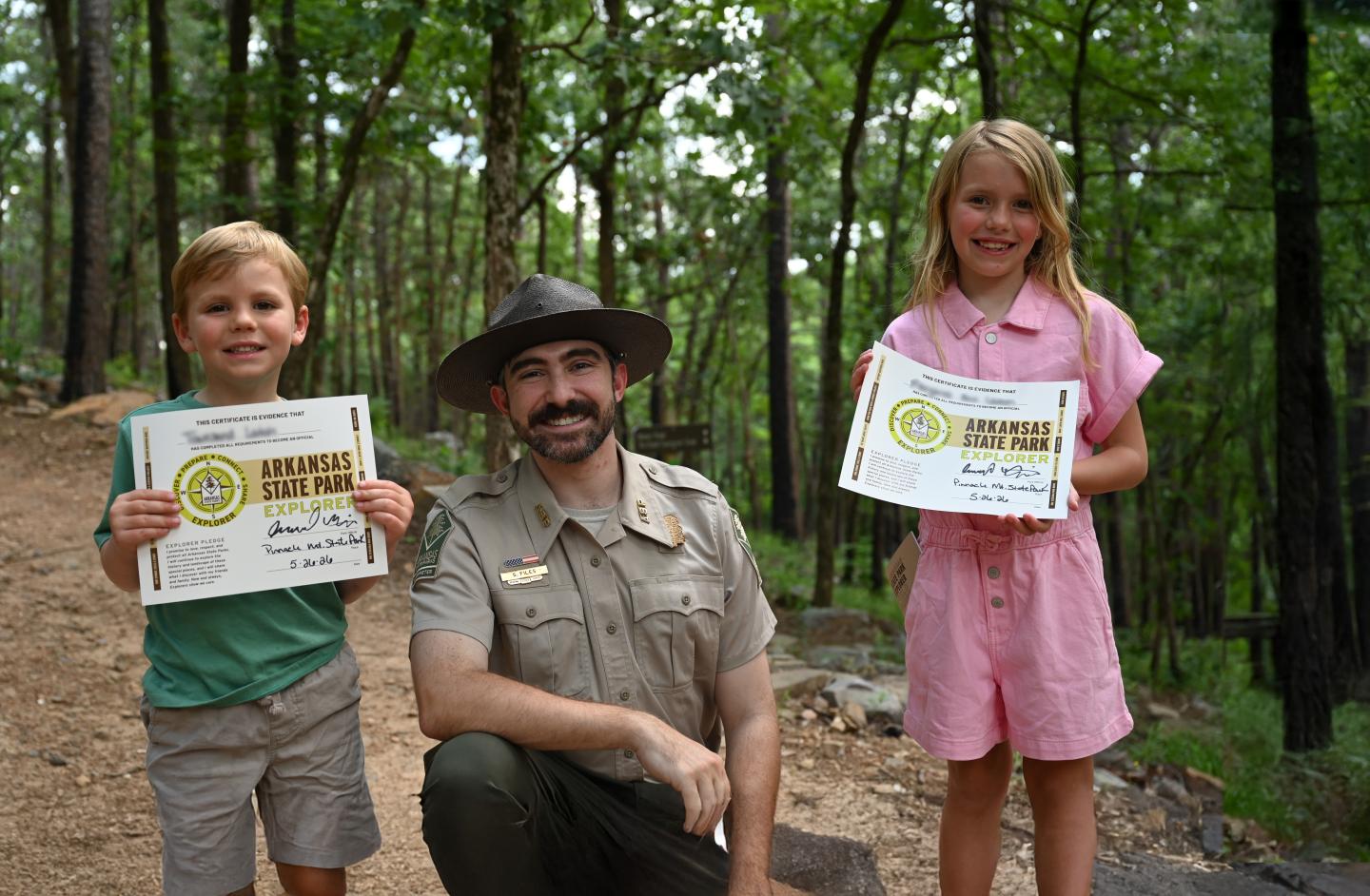 Ranger with two children holding certificates in a forest setting.