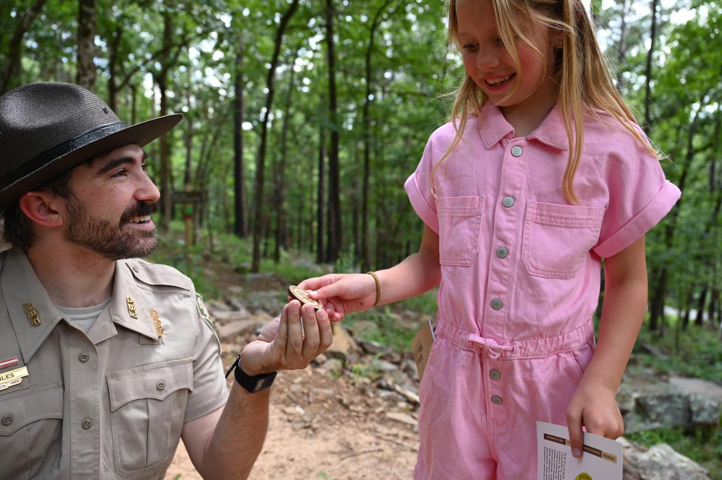 Park ranger smiles, handing badge to girl in pink jumpsuit in a wooded area.