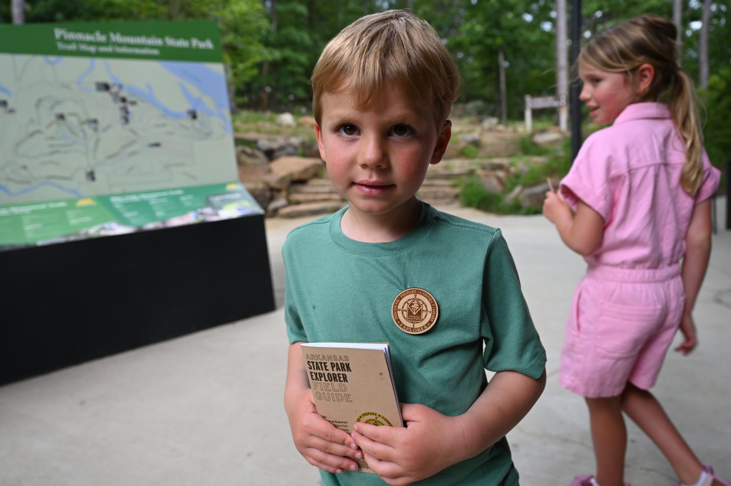 Young boy in green shirt with Explorer badge, holding Explorer booklet, standing outdoors.
