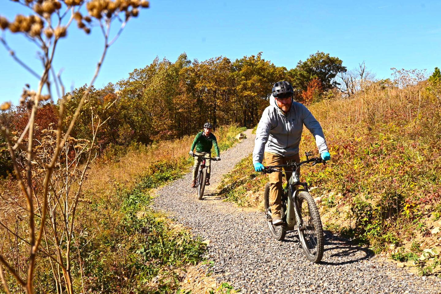 Two cyclists ride on a gravel path through a sunny, autumn landscape.