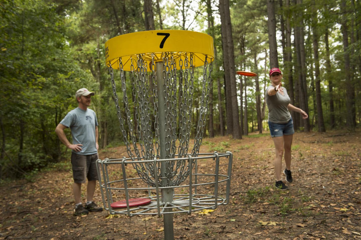 Disc golf game in a forest, woman throws, man watches.