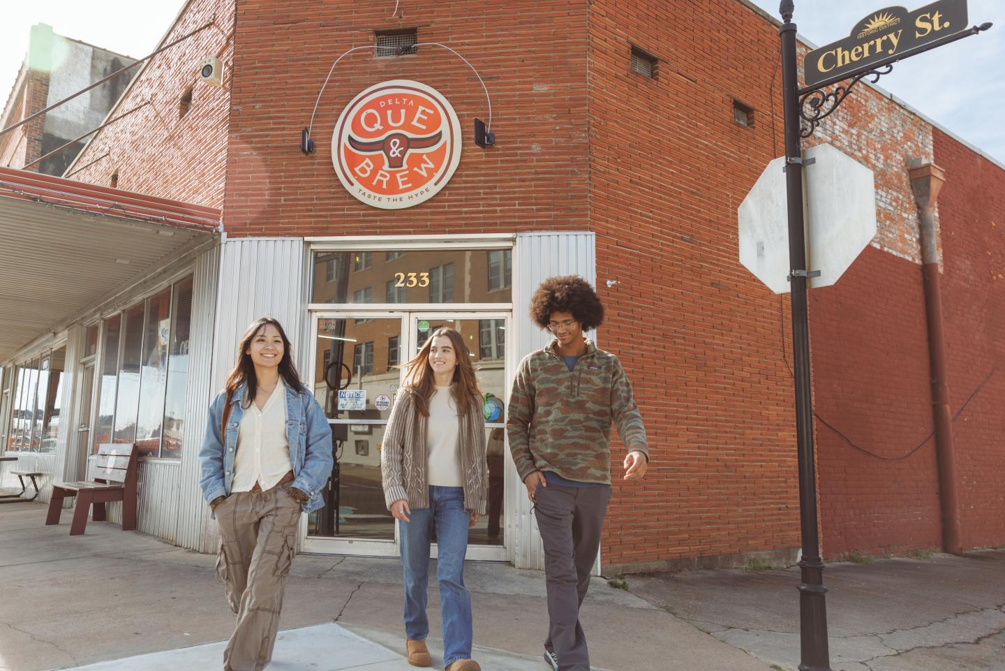 Three people walking past a rustic red brick building on a sunny day.