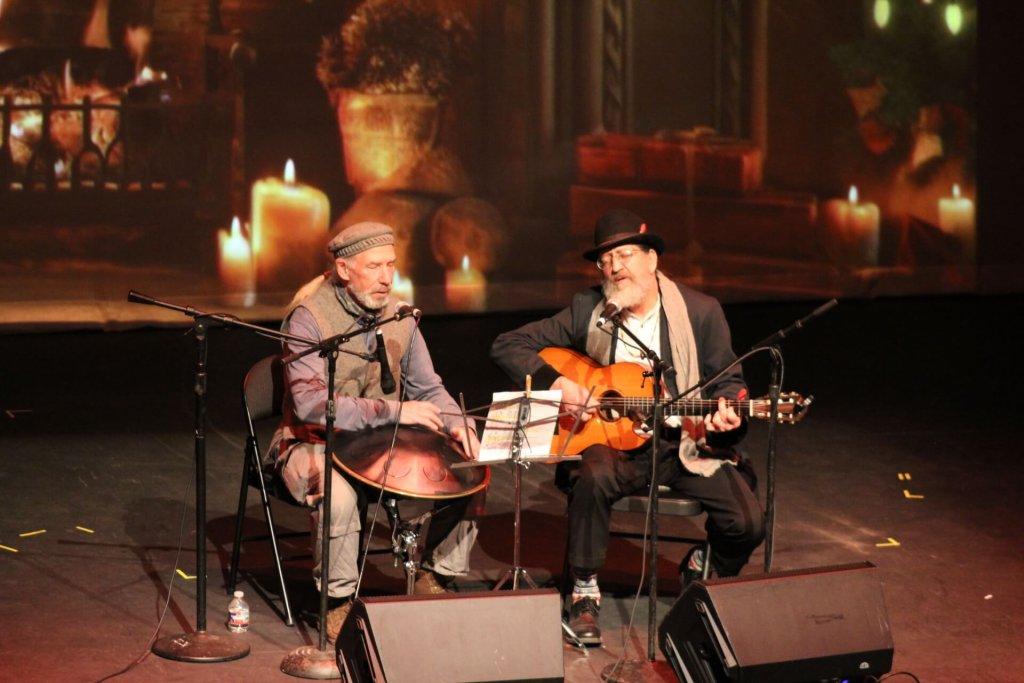 Two musicians on stage with a guitar, surrounded by warm candlelight.