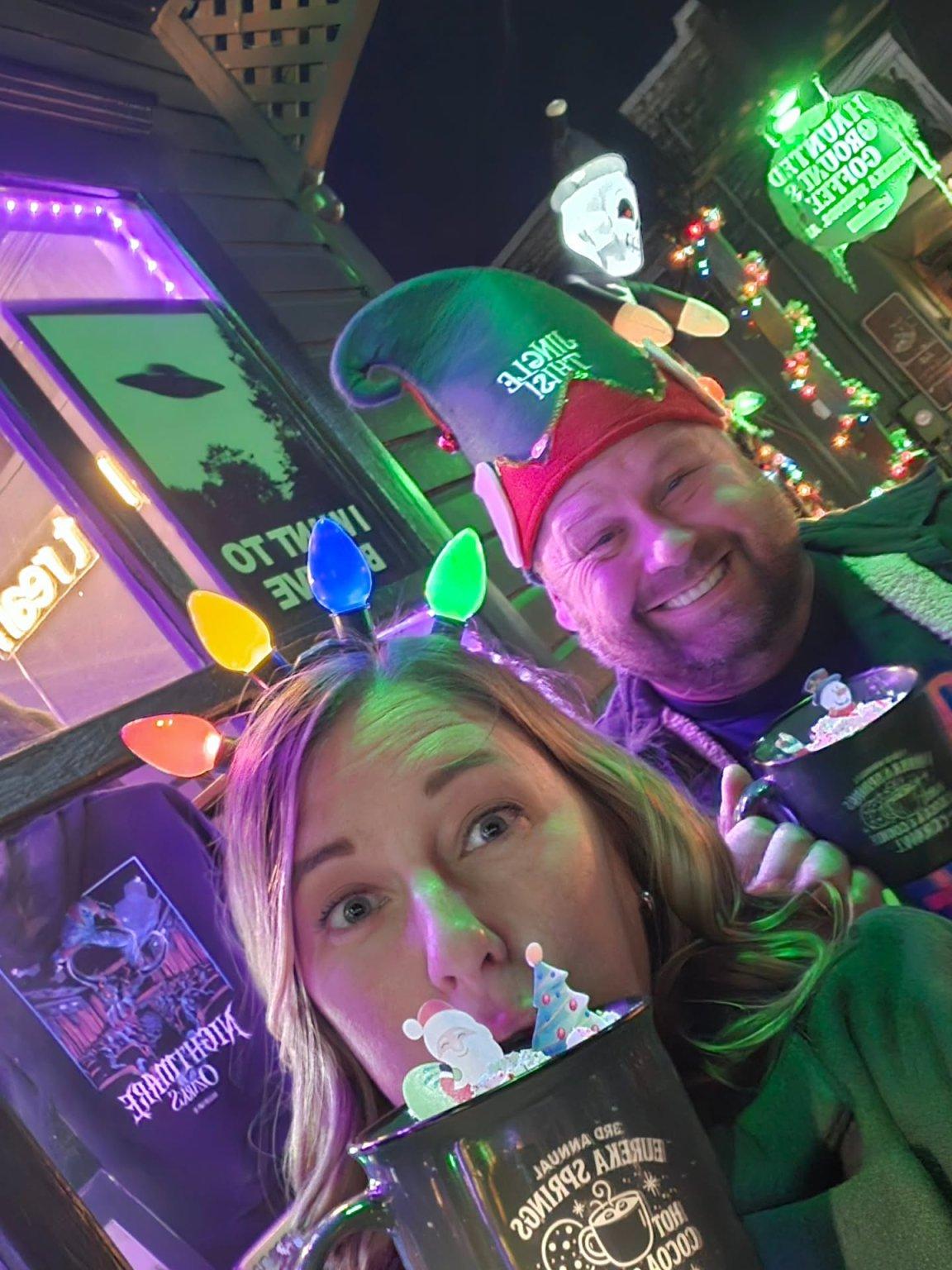 Two people in colorful hats holding mugs, with festive lights in the background.