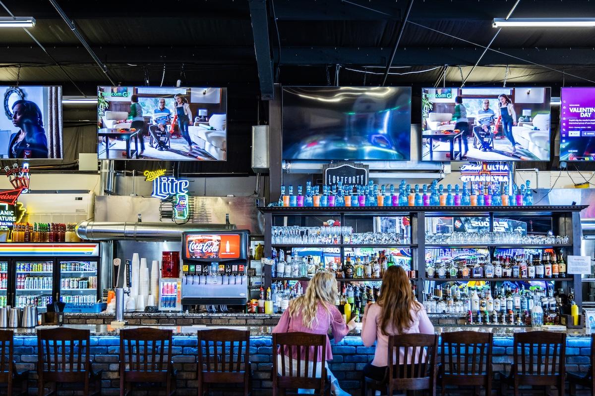Bar interior with two patrons sitting, multiple TVs displaying images above the bar.