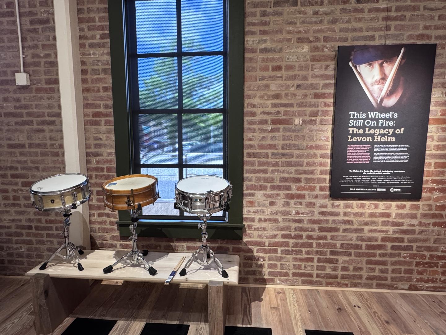 Three snare drums on stands, brick wall and poster in the background.