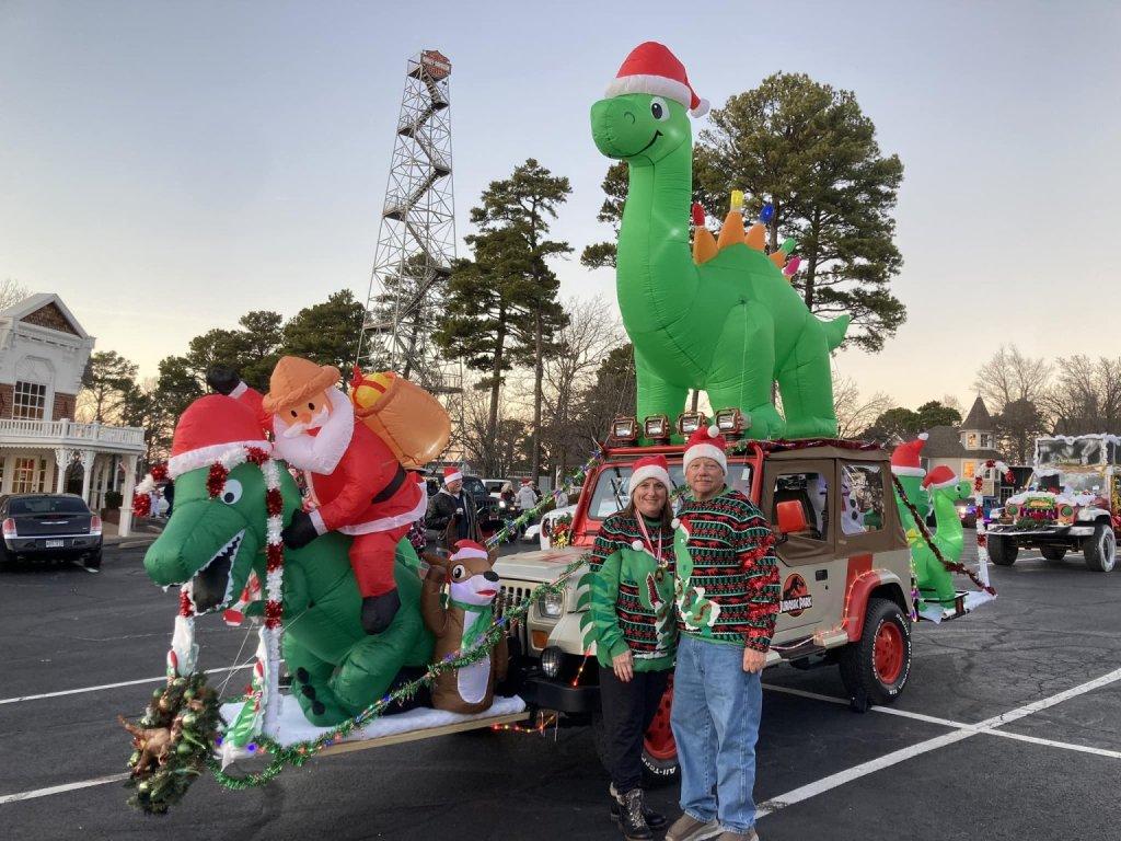Jeep adorned with inflatable dinosaurs, Santa hats. Two people smiling in festive attire.