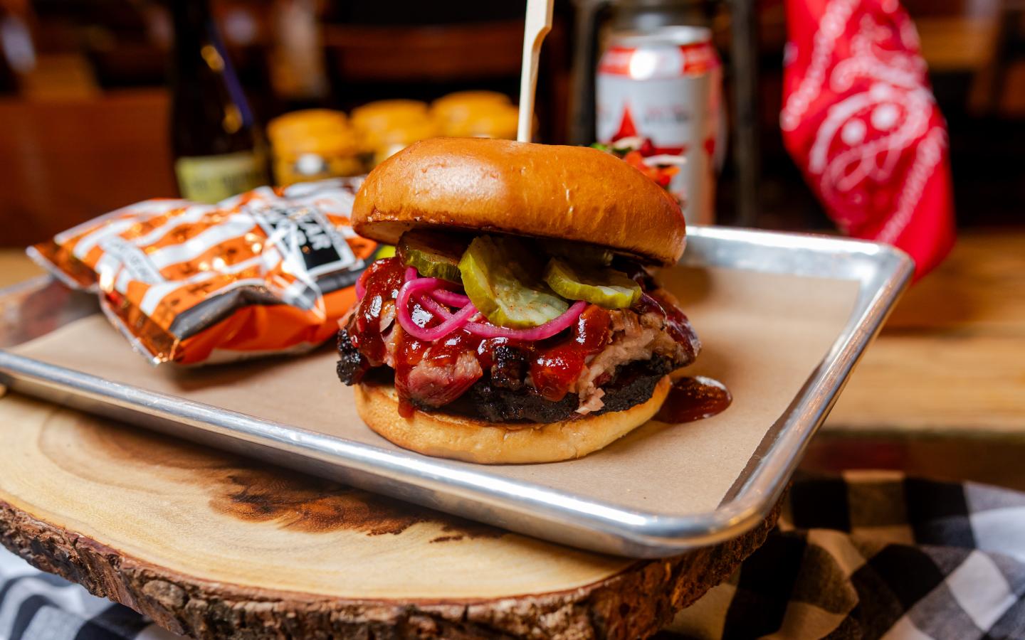 Brisket sandwich with pickles and onions on a tray, chips and drink in the background.