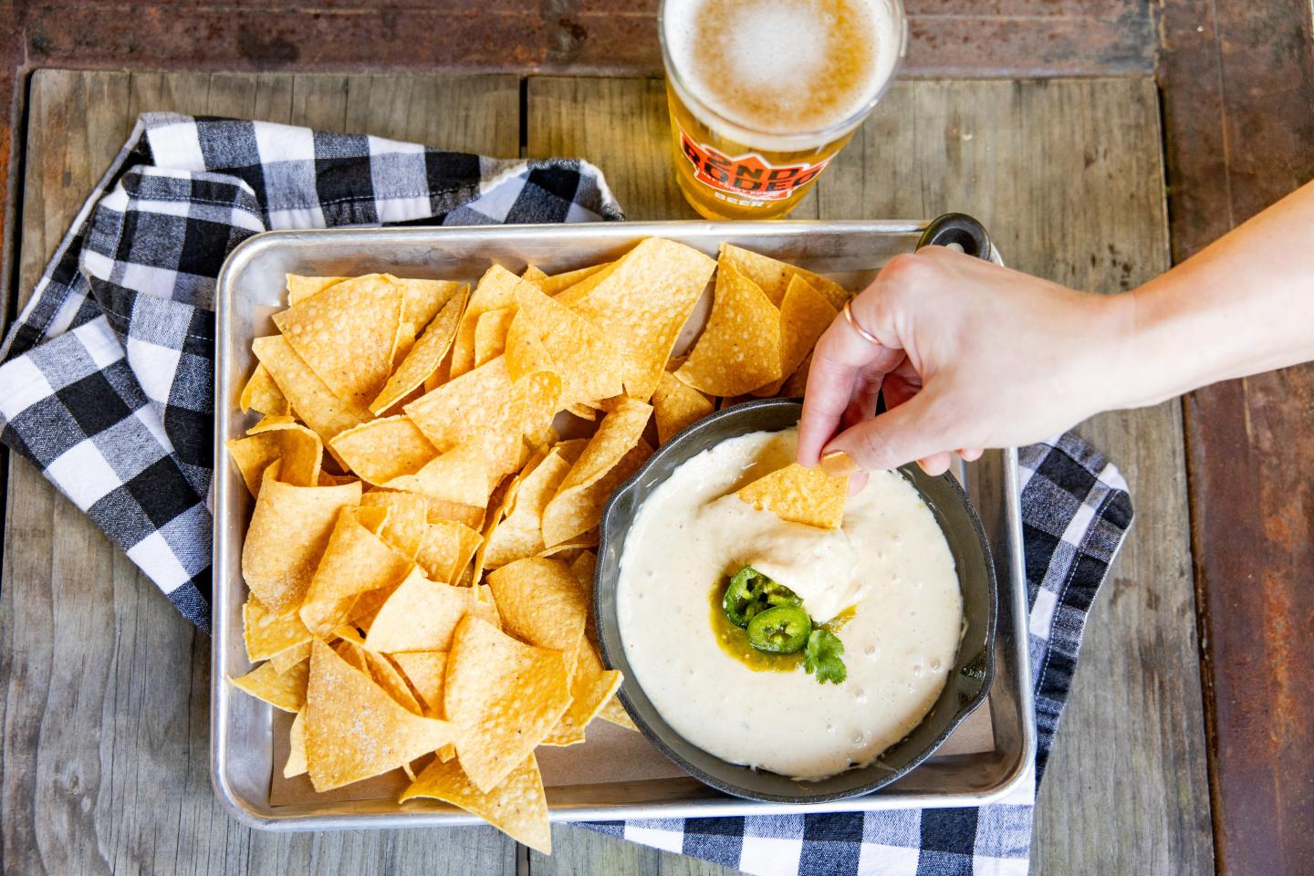 Chips with cheese dip and a hand dipping, next to a glass of beer.