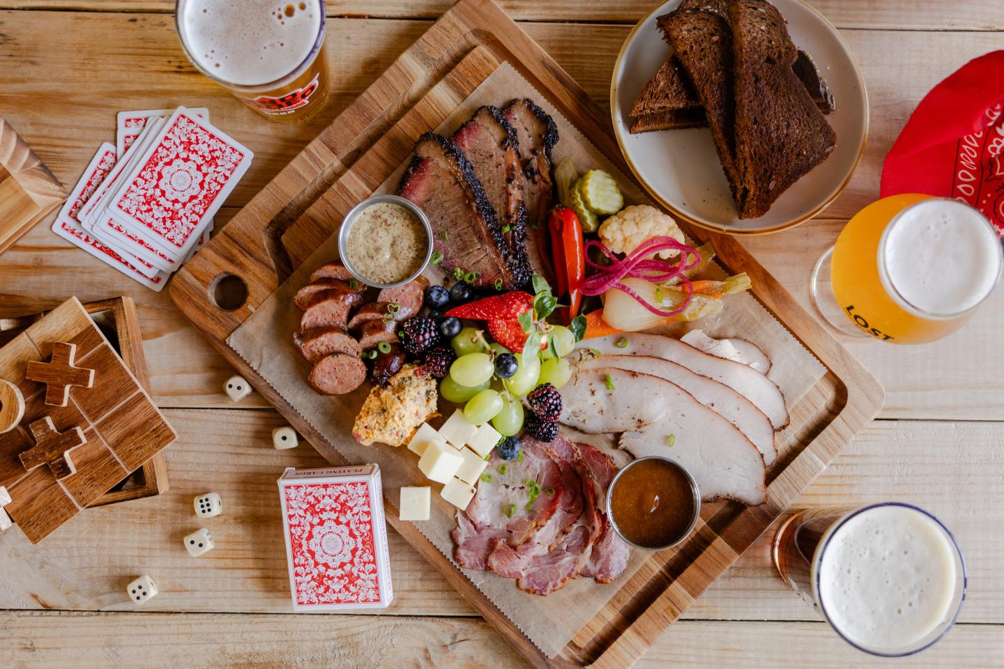 Charcuterie board with meats, cheese, olives, and bread, surrounded by drinks and playing cards.